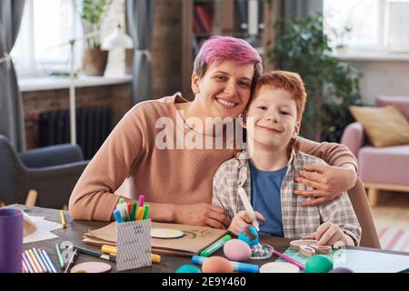 Portrait of happy mother embracing her son and smiling at camera while painting Easter eggs at home Stock Photo