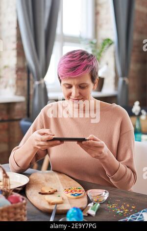 Young woman sitting at the table and making photo of decorated cookies on her mobile phone Stock Photo