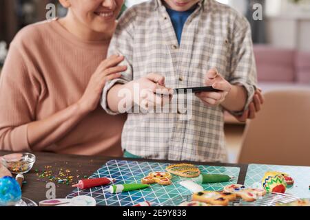 Close-up of boy using mobile phone to make photo of homemade cookies on the table with his mother Stock Photo