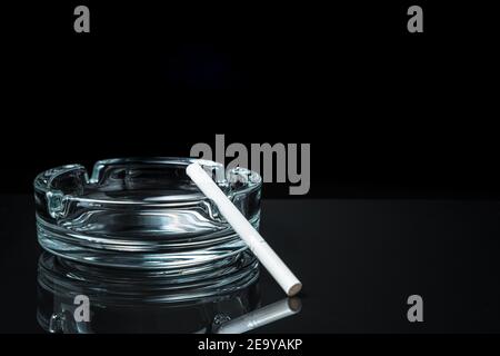 A white cigarette on a transparent ashtray on a black background. Stock Photo