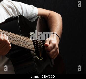 Entertainment concept. Young man playing acoustic guitar, dark background. Guitarist in casual white shirt holds electric black instrument at studio. Stock Photo