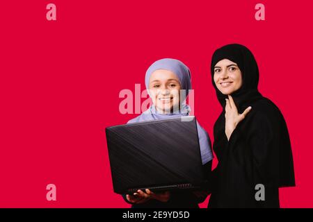 Two muslim female friends standing over red background with a laptop in hands. Young woman in with a covered heads looking to the camera and smiling  Stock Photo
