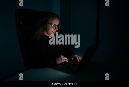 Smiling woman with wireless headphones using a laptop at night. Student working late. Stock Photo