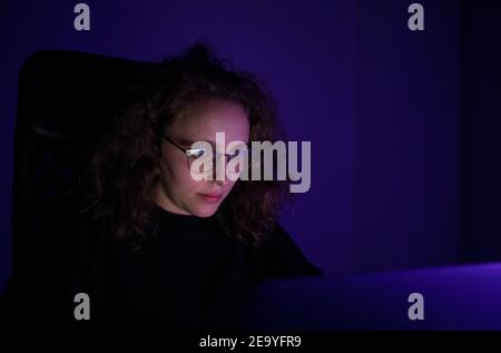 Blond woman with curly hair working late at night on her computer. The ambient light is purple. Overwork concept. Stock Photo