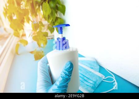 a hand in a medical glove holds a bottle with an antiseptic on a background of green sheets and a white background with masks, hygiene product, protec Stock Photo