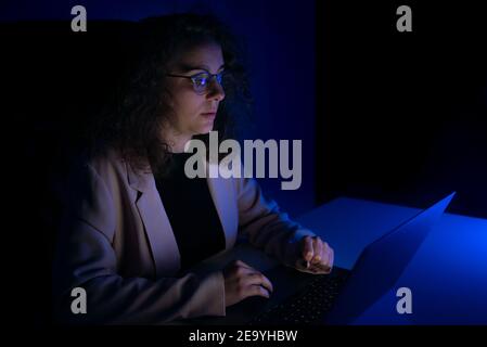 Young woman typing on computer while it is night time. It is dark and there are blue lights. Stock Photo