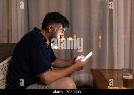 bearded man on sofa uses digital tablet in dining room overnight Stock Photo