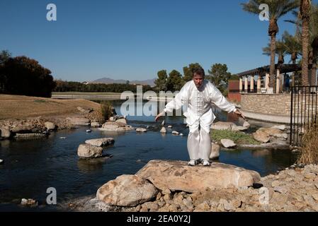 Man wearing a white suit and performs Tai Chi on a rock with water behind him and mountains in background. Stock Photo