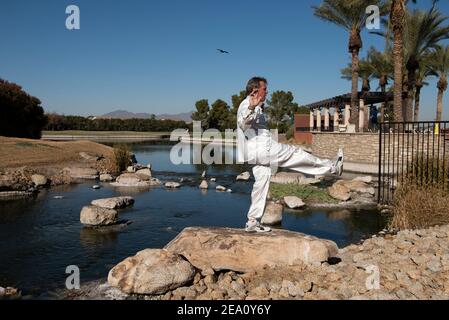 Man wearing a white suit and performs Tai Chi on a rock with water behind him and mountains in background. Stock Photo