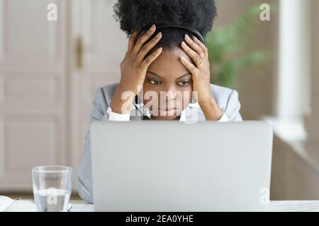 Tired annoyed afro woman wear headphones talking to a bad client looking at screen laptop in office Stock Photo