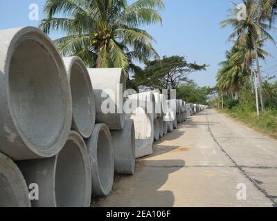 Concrete sewer pipes are stacked at construction site on roadside in Thailand, Road with coconut palm trees and blue sky in background Stock Photo