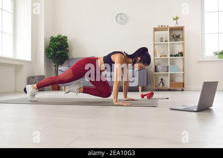 Side view of athletic woman training on a mat in front of a laptop at home in the living room. Stock Photo