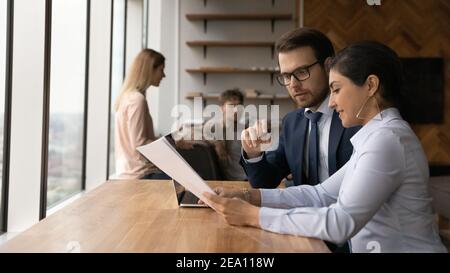 Two diverse teammates mentor intern discuss documents at coworking area Stock Photo