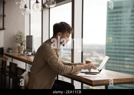 Concentrated millennial man using laptop at desk near panoramic window Stock Photo