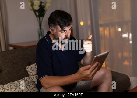 bearded man on sofa uses digital tablet in dining room overnight Stock Photo