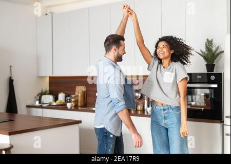 Happy young multi ethnic couple in love dancing and holding hands in the kitchen. Joyful boyfriend and girlfriend have a fun at home, newlywed moved Stock Photo