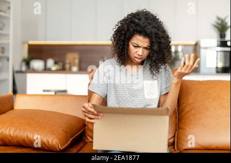 Disappointed African-American woman looking Inside cardboard box, got a wrong online order. Sad biracial girl dissatisfied with parcel Stock Photo