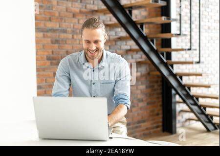 Smiling hipster guy is typing on the laptop, answering emails, chatting in the social networks. Portrait of cheerful young businessman wearing smart Stock Photo