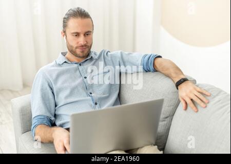 Handsome guy spends time in networks, a young man lying on the comfortable couch and using a laptop, watching movies, working remotely from home Stock Photo