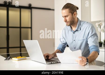 Concentrated guy with a laptop is working on the project remotely, young entrepreneur looks through the work papers, analyzing, checking datas. A male Stock Photo