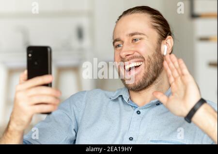 Smiling guy speaking online via video call on the phone, a hipster holds smartphone, looking at screen, waving hello, greeting on video conference Stock Photo