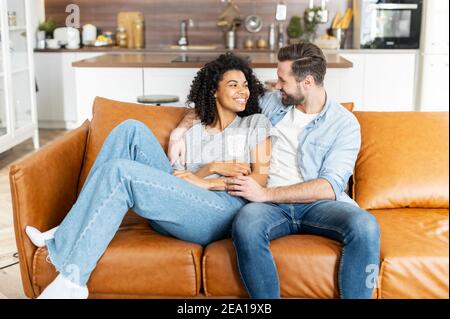 Lovely multiracial couple in embraces in cozy living room at home. Hilarious interracial couple enjoys time together at home sitting on the Stock Photo