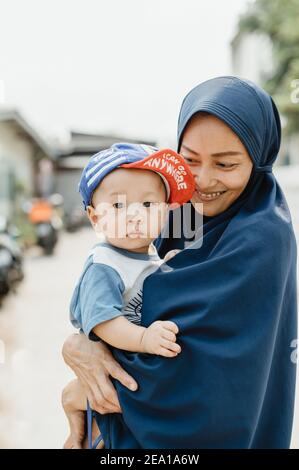 Thai Muslim woman wearing a blue hijab holding a baby with a cap, smiling and looking at him with affection. Stock Photo
