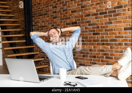 Cheerful young businessman leaned back in the chair, holds his hands behind his head and looks at laptop. Smiling freelancer feeling satisfaction Stock Photo