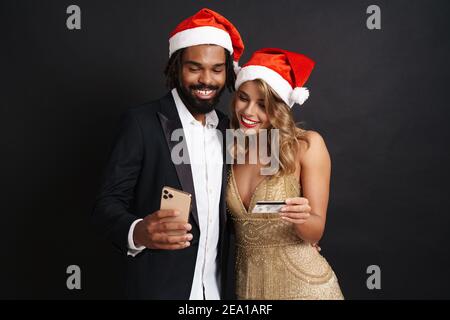 Happy young couple wearing Christmas hats using mobile phone hold credit bank card isolated on black background. Happy New Year celebration merry holi Stock Photo