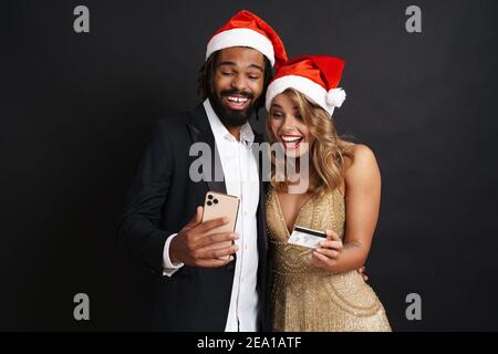Happy young couple wearing Christmas hats using mobile phone hold credit bank card isolated on black background. Happy New Year celebration merry holi Stock Photo
