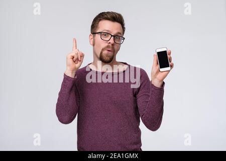Cheerful, positive guy having smart phone with black screen in hand, pointing with forefinger up giving advice. Studio shot Stock Photo