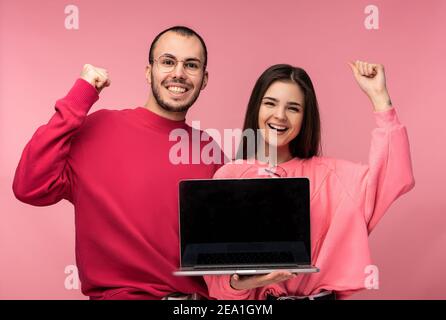 Photo of sweet couple holds laptop. Male and female look blessed and smile, isolated over pink background Stock Photo