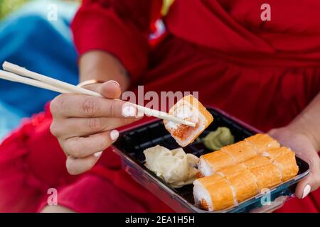 Close-up sushi set. Attractive girl in red dress holds sushi with chopsticks in hands. Courier delivered sushi set for pretty woman. Black box with su Stock Photo