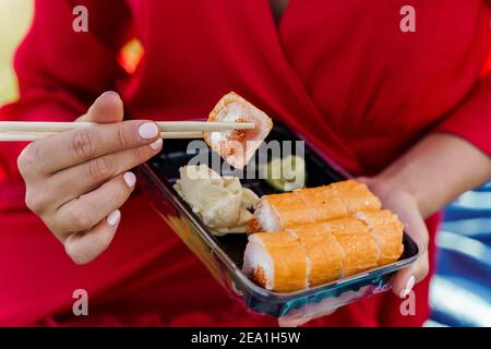 Close-up sushi set. Attractive girl in red dress holds sushi with chopsticks in hands. Courier delivered sushi set for pretty woman. Black box with su Stock Photo