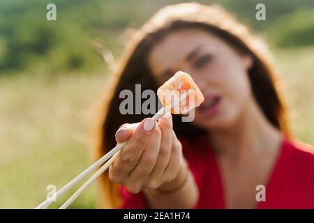 Close-up one sushi. Attractive girl holds sushi with chopsticks in hands and gives to you. Courier delivered sushi set for pretty woman. Black box wit Stock Photo