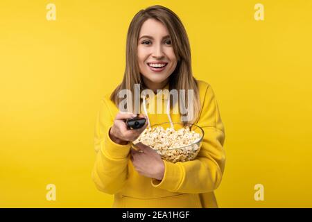 Photo of attractive lady holds tv remote and plate of popcorn, with happy face tries to turn a channel on. Wears casual yellow hoody, isolated yellow Stock Photo
