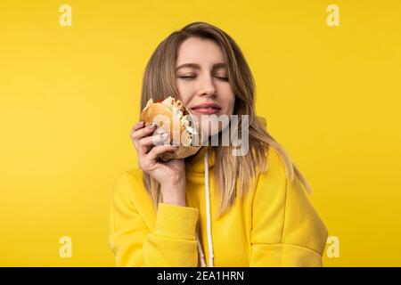 Photo of attractive lady poses in a cute way with burger. Wears casual yellow hoody, isolated yellow color background Stock Photo