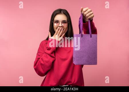 Photo of attractive lady in glasses holds present, surprised and happy. Wears casual pink sweater white pants isolated pink color background Stock Photo