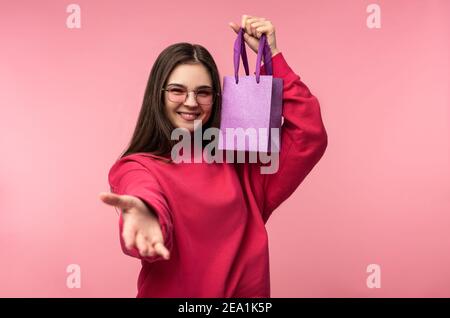 Photo of attractive lady in glasses holds present, pleasant lend a hand, wears casual pink sweater white pants isolated pink color background Stock Photo