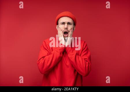 Photo of attractive man with beard in glasses and red clothing. Male shocked and mouth opened, isolated over red background Stock Photo
