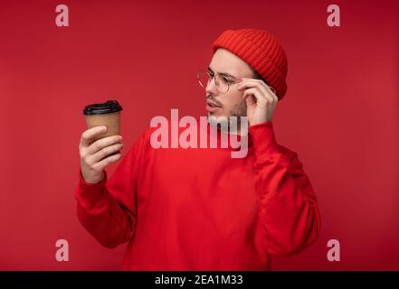 Photo of attractive man with beard in glasses and red clothing. Curious man looks at his coffee cup, isolated over red background Stock Photo