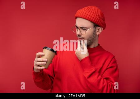 Photo of attractive man with beard in glasses and red clothing. Curious man looks at his coffee cup, isolated over red background Stock Photo