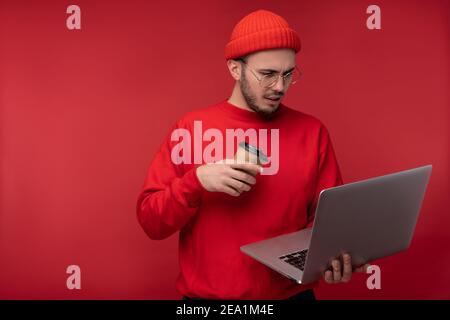 Photo of attractive man with beard in glasses and red clothing. Thoughtful man holds laptop and coffee, isolated over red background Stock Photo