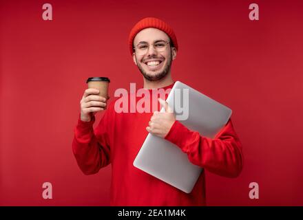 Photo of attractive man with beard in glasses and red clothing. Happy man holds laptop and coffee feels good, isolated over red background Stock Photo