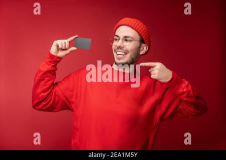 Photo of attractive man with beard in glasses and red clothing. Happy man holds and points at credit card, isolated over red background Stock Photo