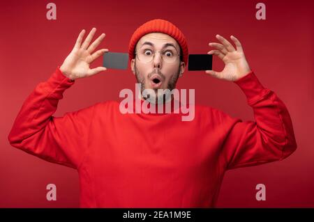 Photo of attractive man with beard in glasses and red clothing. Surprised man holds 2 credit cards, isolated over red background Stock Photo