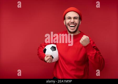 Photo of attractive man with beard in glasses and red clothing. Happy man holds ball and smiles, isolated over red background Stock Photo