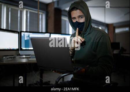 Male hacker in hood works on laptop in dark office Stock Photo
