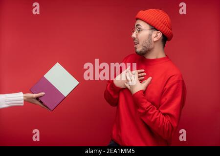 Photo of surprised man with beard in glasses and red clothing. Male unexpectedly gets gift, isolated over red background Stock Photo