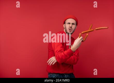 Photo of happy man with beard in glasses and red clothing. Plays sausages like nunchucks ninja, isolated over red background. Stock Photo